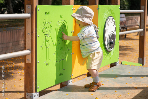 Happy emotional baby boy wearing summer clothes explores a playground. He is interacting with a green activity panel featuring simple drawings of a boy and girl designed for sensory play. 
