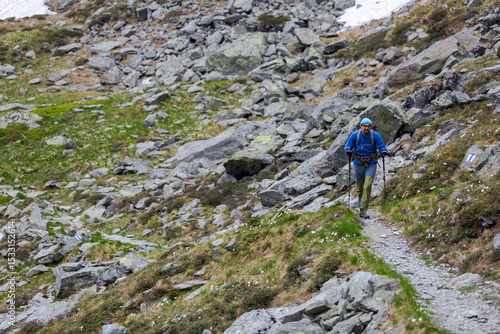 Mountain hiker with backpack and trekking poles navigating rocky alpine trail through rugged terrain filled with boulders and scattered stones