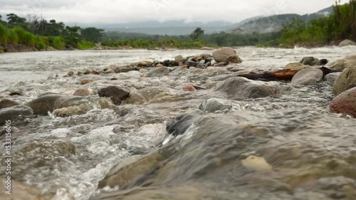 Crystal clear water flows over rocks in a river in the Amazon rainforest, with a cloudy sky and lush vegetation in the background