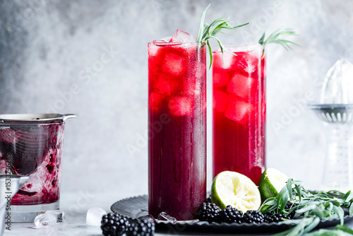 Two tall glasses of refreshing berry cocktail with ice, garnished with herbs and served with blackberries and lime halves on a dark plate.