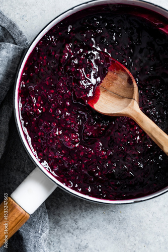 A saucepan filled with dark berry jam being stirred with a wooden spoon on a gray surface.