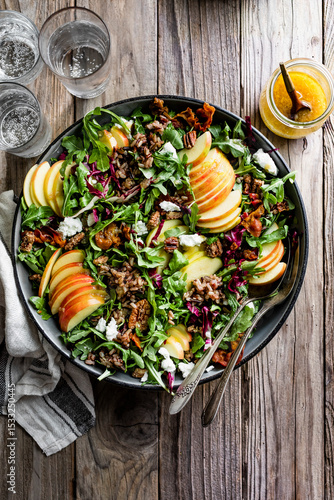 A fresh salad with arugula, apple slices, goat cheese, pecans, and grains served on a wooden table with a bowl of dressing and glasses of water.
