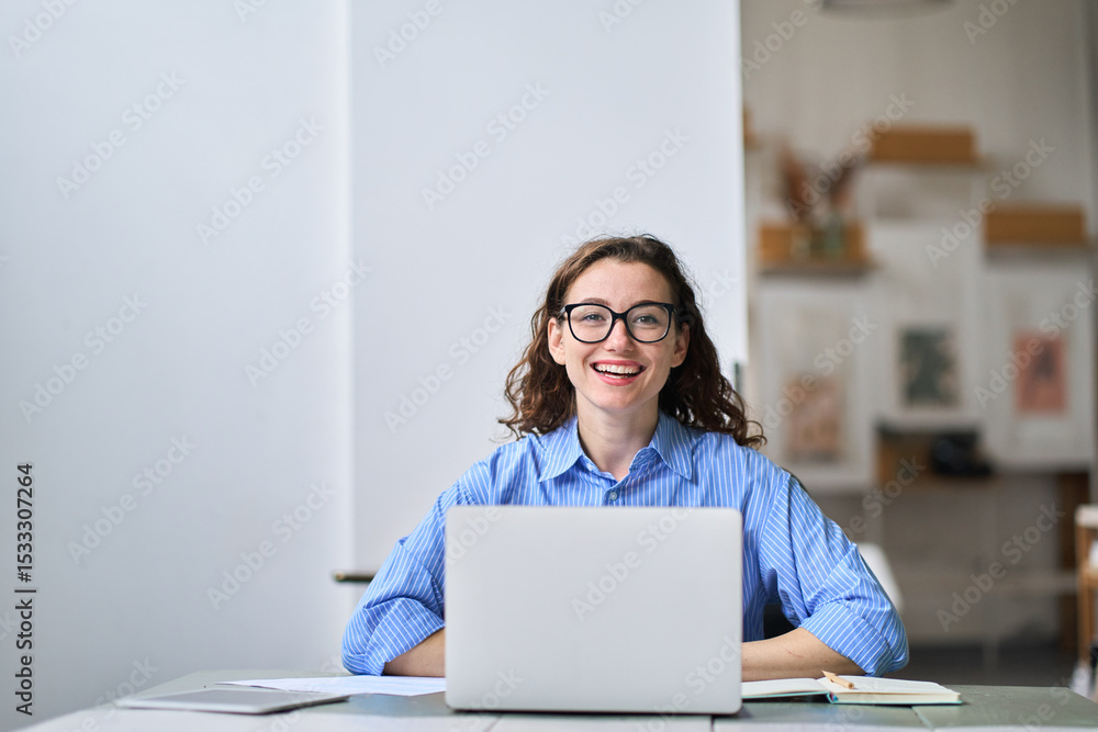 Obraz premium Portrait of young happy business woman company employee sitting at desk working on laptop. Smiling female professional designer or student using computer in corporate office looking at camera.