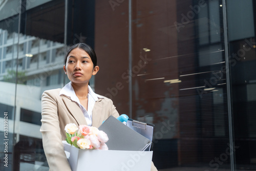 Young asian woman in formal suit carrying a box of personal belongings after being laid off, looking sad and disappointed while walking outside office building, job loss, unemployment crisis.