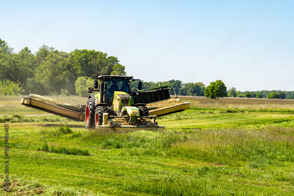 Fototapeta premium Tractor with wide mower cutting grass on green field