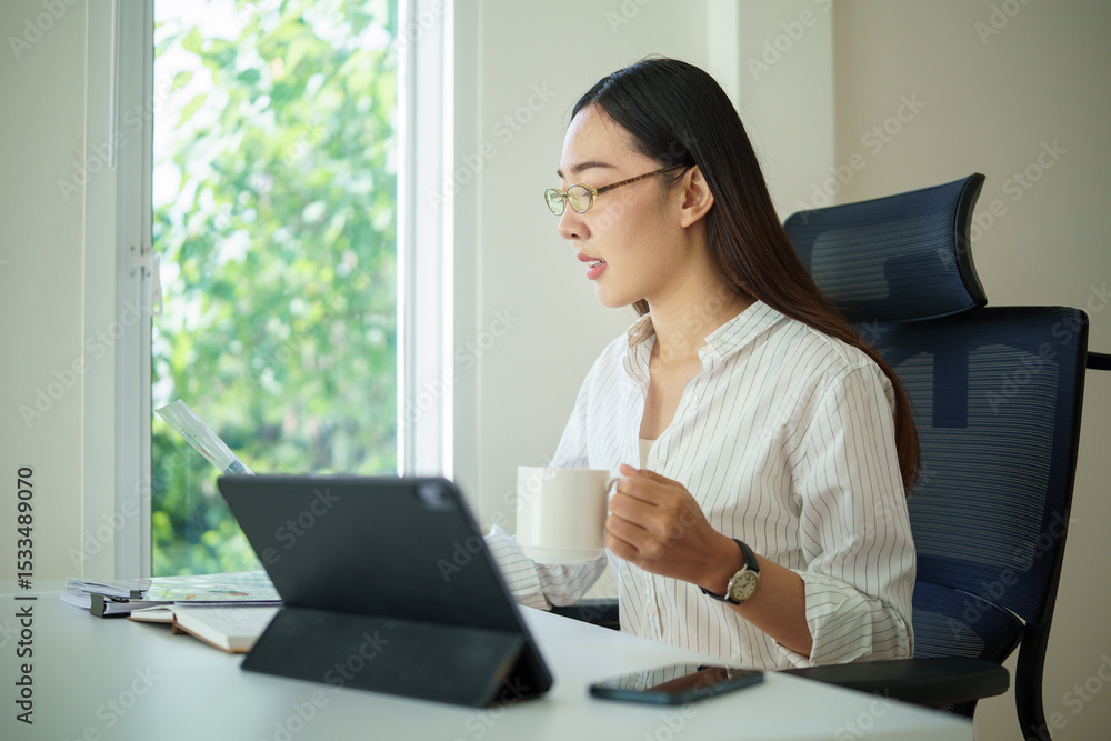 Young businesswoman holding a cup while analyzing reports on a tablet near a bright office window. Young businesswoman holding a cup while analyzing reports on a tablet near a bright office window.