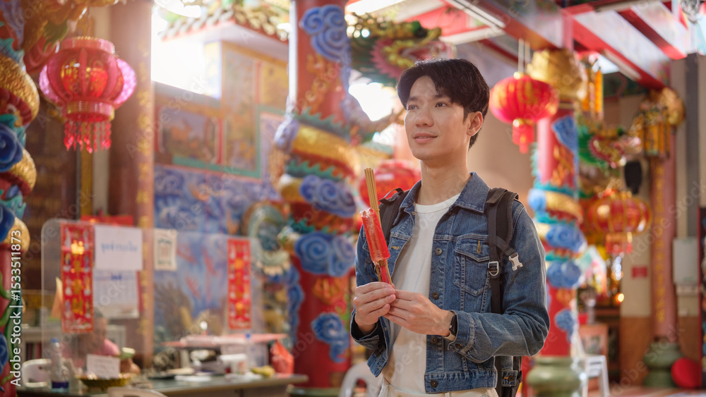 Obraz premium Young male traveler holding incense sticks while making a wish inside a Chinese temple.
