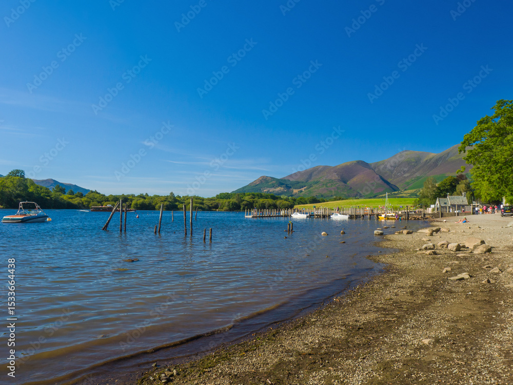Obraz premium Calm Derwentwater lakeside with a jetty, grassy park and mountains in the distance (Keswick, Lake District, England, United Kingdom)
