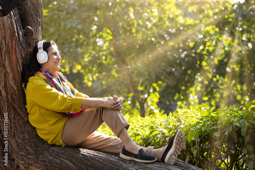 Canvas Print Woman listening to music on headphones while sitting on a tree trunk in the garden