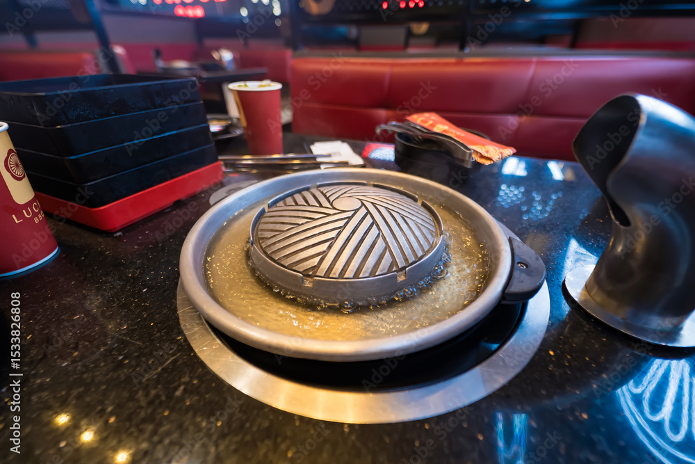 Obraz premium Korean BBQ grill pan on a table in a restaurant with metal grill and broth around, ready for cooking with condiments nearby