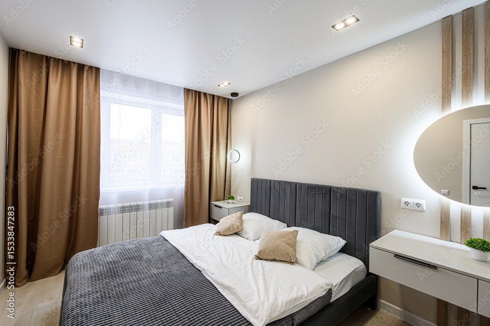Fototapeta premium modern bedroom featuring a bed with a gray headboard, white linens, brown curtains, and a round, illuminated mirror