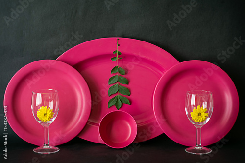 Image of a dinner set against dark background shot in window light. Pink Set of oval plates, round plates, bowl and some curry leaves along with yellow flower for decoration.