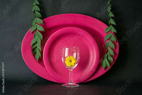 Image of a pink dinner set against dark background shot in window light. Oval and round plates set with curry leaves and wine glass with yellow flower.