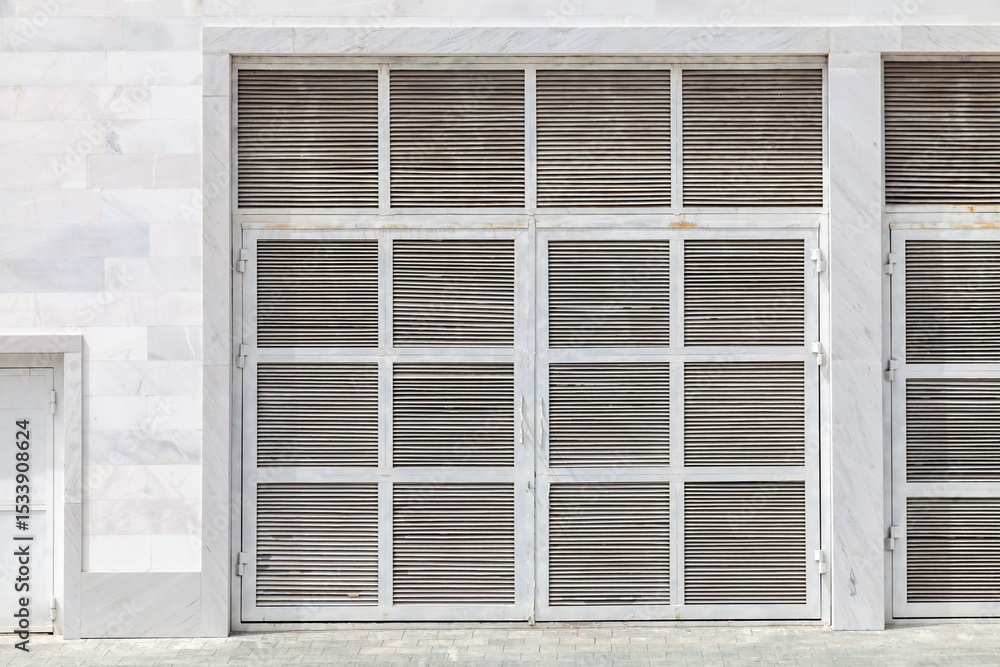 Fototapeta premium A concrete utility storage unit with two white doors is placed in a quiet outdoor area, surrounded by greenery and fallen leaves, during daytime.