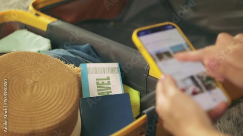 Traveler booking hotel online using smartphone sit at home beside packed suitcase, passport, ticket, clothes, straw hat displayed.Traveling and vacation concept