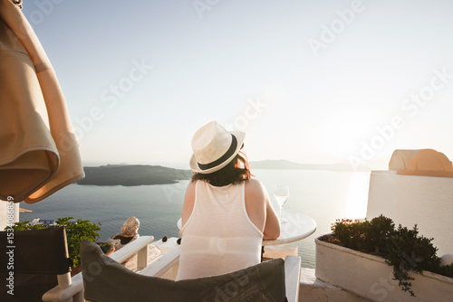 A woman enjoys a scenic sunset view over the Aegean Sea from a Santorini terrace with a glass of wine and a vape.