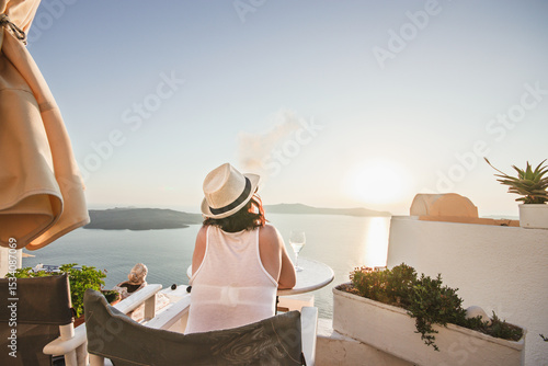 A woman enjoys a scenic sunset view over the Aegean Sea from a Santorini terrace with a glass of wine and a vape.