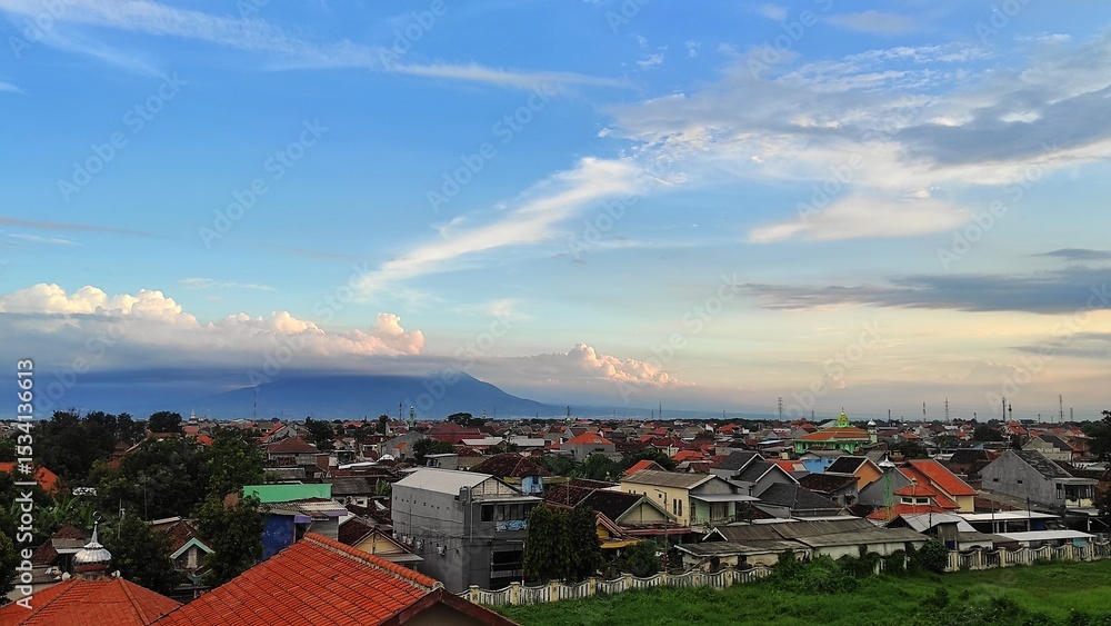 Fototapeta premium Wide-angle cityscape with rows of rooftops in the foreground, a towering volcano partially obscured by clouds in the background, captured during golden hour with soft light and pastel sky tones