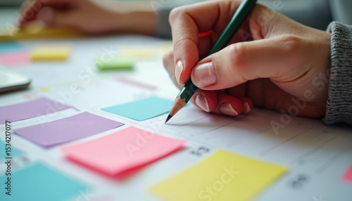 Woman planning schedule using colourful paper calendar with pencil in office. Time management, note taking, planning workflow, organisation concept. Female hand writes schedule on colorful sticky