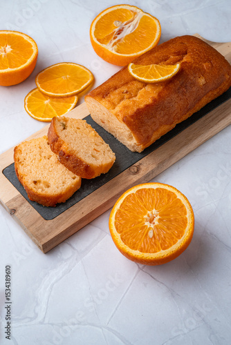 Orange pound cake with slices of fresh orange on wooden board. Sweet loaf cut into pieces, citrus fruits arranged on white marble background. Minimal food styling and dessert composition.