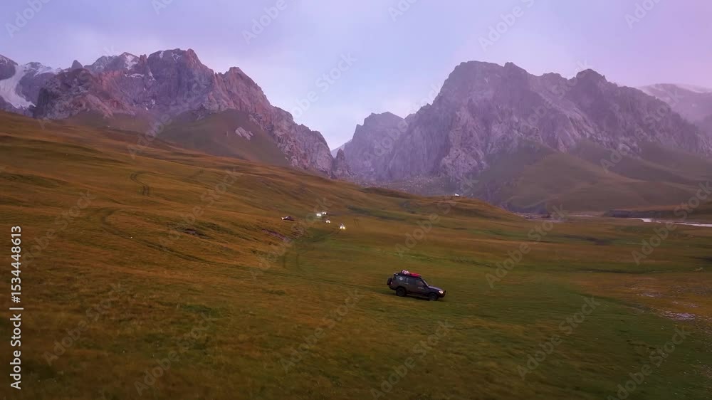 An off-road vehicle drives across a grassy mountain valley with dramatic peaks in the distance, under the soft light of dusk or dawn An off-road vehicle drives across a grassy mountain valley with dramatic peaks in the distance, under the soft light of dusk or dawn