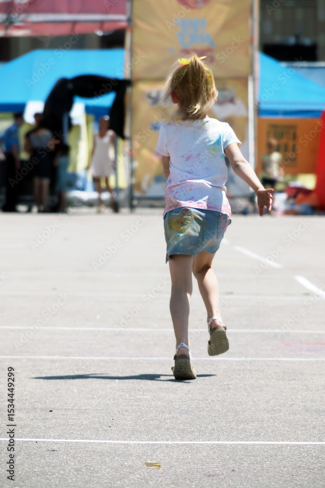 Fototapeta premium A little girl runs and skips across the asphalt towards a some woman during the Holi festival celebration. On the back of her skirt you can see a yellow and blue heart with red spots.