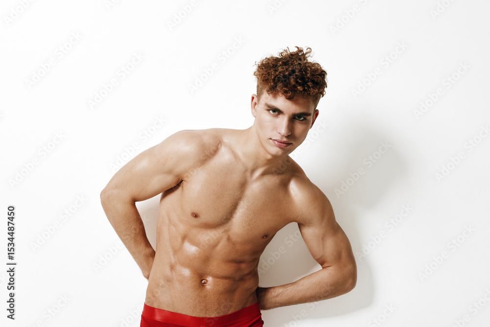 Confident young man with curly hair posing against a clean white background, showcasing a toned physique in red swimwear. Fitness and health concept. Confident young man with curly hair posing against a clean white background, showcasing a toned physique in red swimwear. Fitness and health concept.