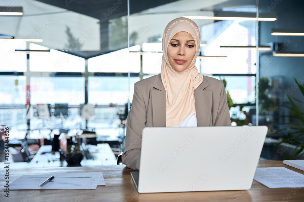 Obraz premium Focused professional finance specialist business lady wearing hijab working on laptop pc sitting at desk in office. Young adult middle eastern muslim woman using computer for work doing banking online