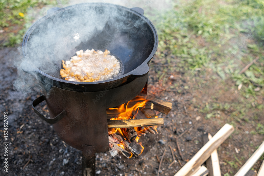 Cooking Traditional Dish Over Open Fire With Cast Iron Pot in a Rural Outdoor Setting During Daylight Cooking Traditional Dish Over Open Fire With Cast Iron Pot in a Rural Outdoor Setting During Daylight