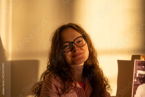 Portrait of a young woman with curly hair and eyeglasses, smiling brightly while basking in the warm, golden hour sunlight