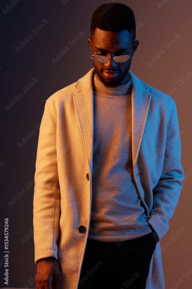 Obraz premium Stylish young man wearing a beige coat and sunglasses, exhibiting a thoughtful emotion against a gradient background
