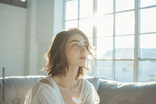 Serene woman bathed in soft sunlight sitting by a large window with natural light filling the space  evoking calm and tranquility