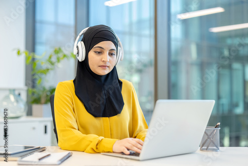 An observant Muslim woman wearing a hijab and headphones is working on a laptop in a modern office setting, possibly attending an online class.