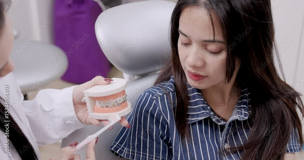 Asian female dentist adult holding dental model with braces and demonstrating brushing technique to smiling young asian female patient during dental hygiene consultation in modern dental clinic Asian female dentist adult holding dental model with braces and demonstrating brushing technique to smiling young asian female patient during dental hygiene consultation in modern dental clinic