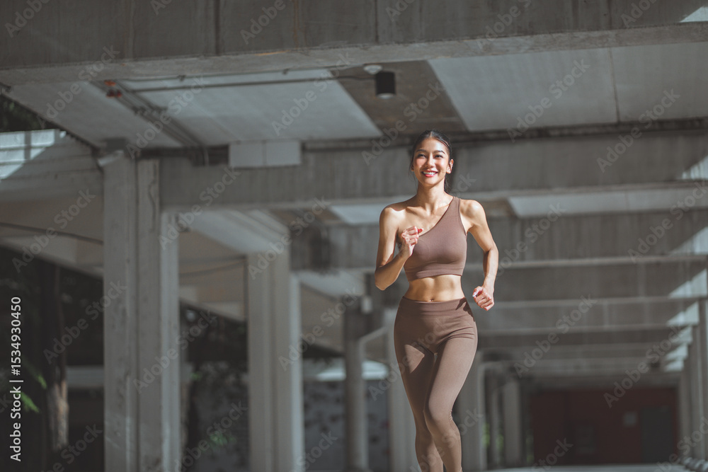 Fototapeta premium Happy asian female athlete wearing activewear running outdoors with concrete city buildings in background – energetic woman embracing urban fitness, active lifestyle, and healthy morning exercise