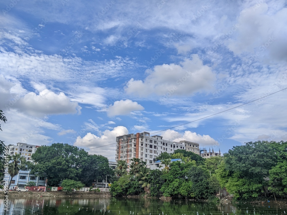 Bright urban landscape with buildings, lush green trees, and a clear blue sky filled with fluffy clouds, reflected on a calm lake. Peaceful city nature scene. Bright urban landscape with buildings, lush green trees, and a clear blue sky filled with fluffy clouds, reflected on a calm lake. Peaceful city nature scene.