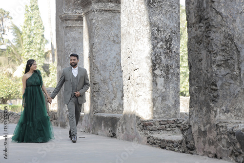 A stylish Indian couple poses affectionately at an ancient stone monument. The woman wears a flowing dark green gown, and the man is dressed in a classic three-piece suit.