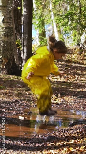 Happy child in yellow rain suit jumping in puddle in park on a sunny day. Active outdoor play, fun and nature exploration in Scandinavian woodland environment.