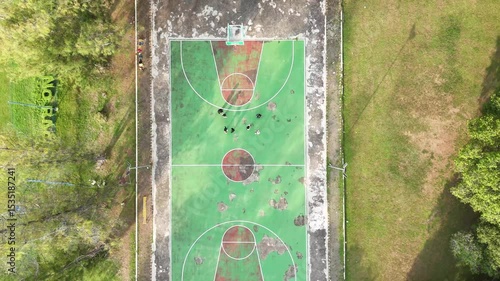 A topdown view shows a worn green  red basketball court surrounded by grass and trees People are playing on the court