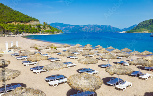 Fototapeta Naklejka Na Ścianę i Meble -  Sandy beach in Icmeler, Turkey, with rows of white sunbeds and straw parasols along turquoise shoreline. Icmeler is popular coastal resort surrounded by green hills and calm sea under clear sky. 