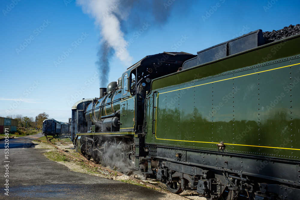 Fototapeta premium Green steam locomotive idles on tracks beside sunlit platform