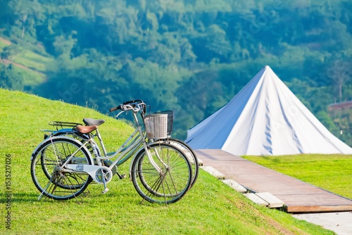 A vintage bicycle with a front basket parked on a green grassy hill near a white tent with a scenic mountain backdrop