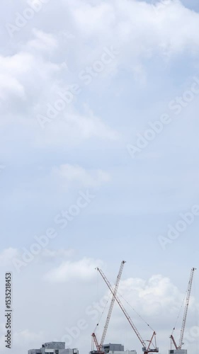 Time lapse of fluffy white clouds moving against blue sky with construction cranes in foreground. Summer nature weather sky. 