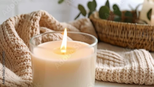 Burning candle in a glass with a beige knitted blanket and plants adds a cozy atmosphere, creating soft light and warm feeling.
