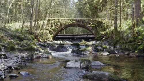 drone flying low over Ognon river with old stone bridge and waterfalls in pine tree forest in Vosges