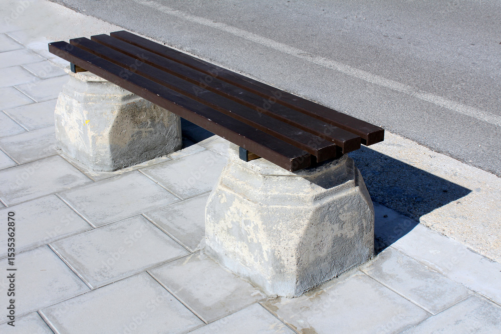 Obraz premium Simple park bench with dark wooden slats and sturdy concrete supports stands on a paved sidewalk by the road, casting a shadow in bright sunlight, offering a resting spot in an urban setting
