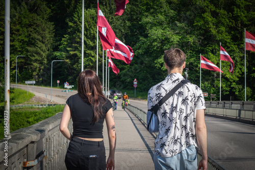 A young man and woman walk across a bridge lined with Latvian flags on a sunny day, surrounded by lush greenery and cyclists approaching in the distance.