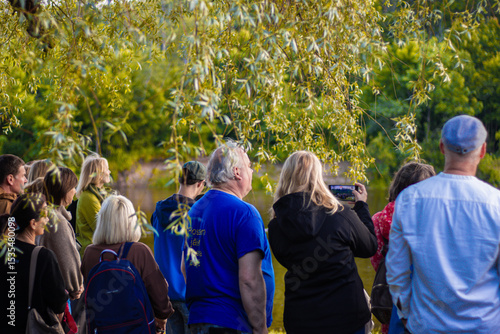 A diverse group of people stands near a calm forest lake, watching an event or view, with one person filming the scene using a smartphone under leafy tree branches.