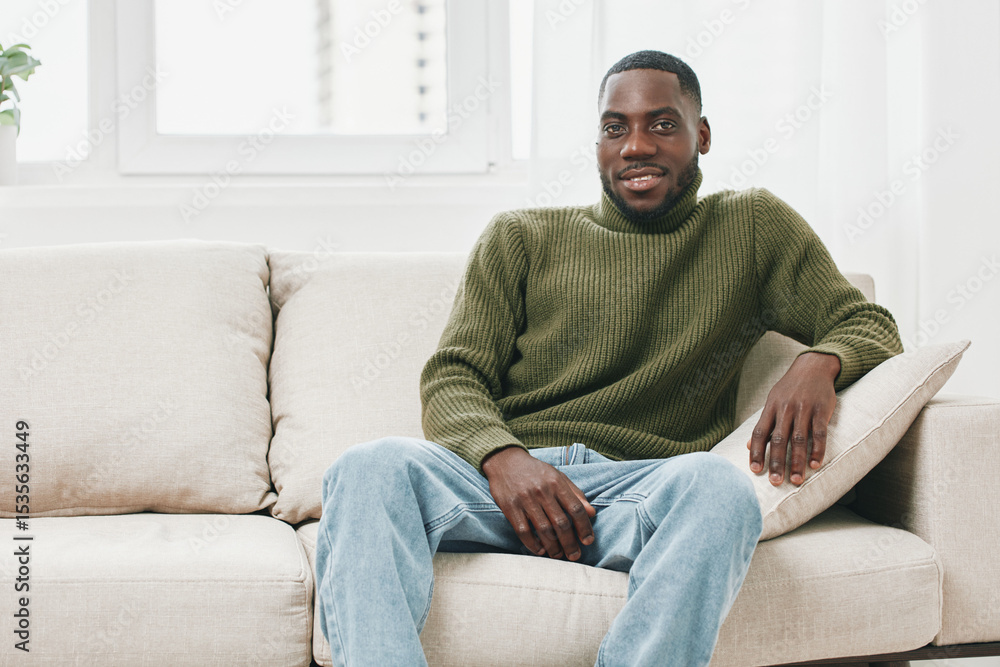 Relaxed young Black man smiling in a cozy living room, wearing a green sweater and blue jeans, representing comfort and happiness at home Relaxed young Black man smiling in a cozy living room, wearing a green sweater and blue jeans, representing comfort and happiness at home