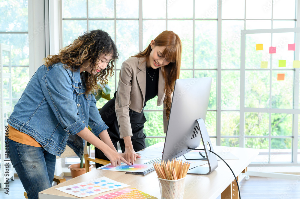 Obraz premium Happy two young female graphic designers discussing with colleague in office. Creative people working together using computer at desk.